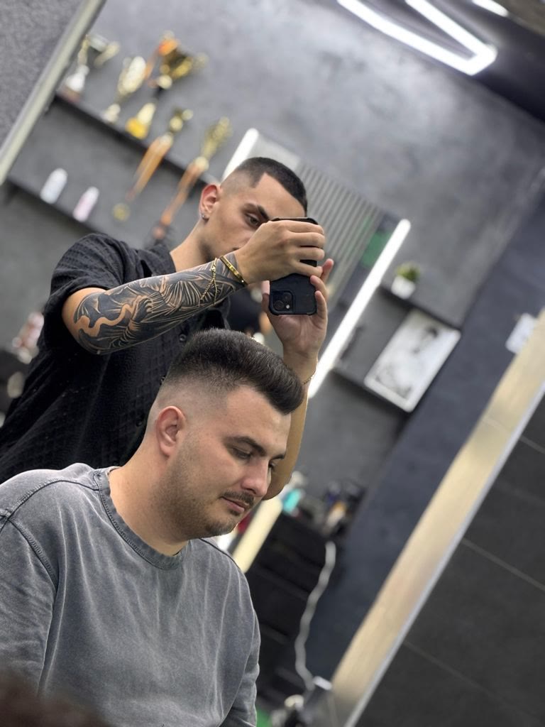 Frizer cu brațul tatuat aranjând părul unui client, trofee pe raft